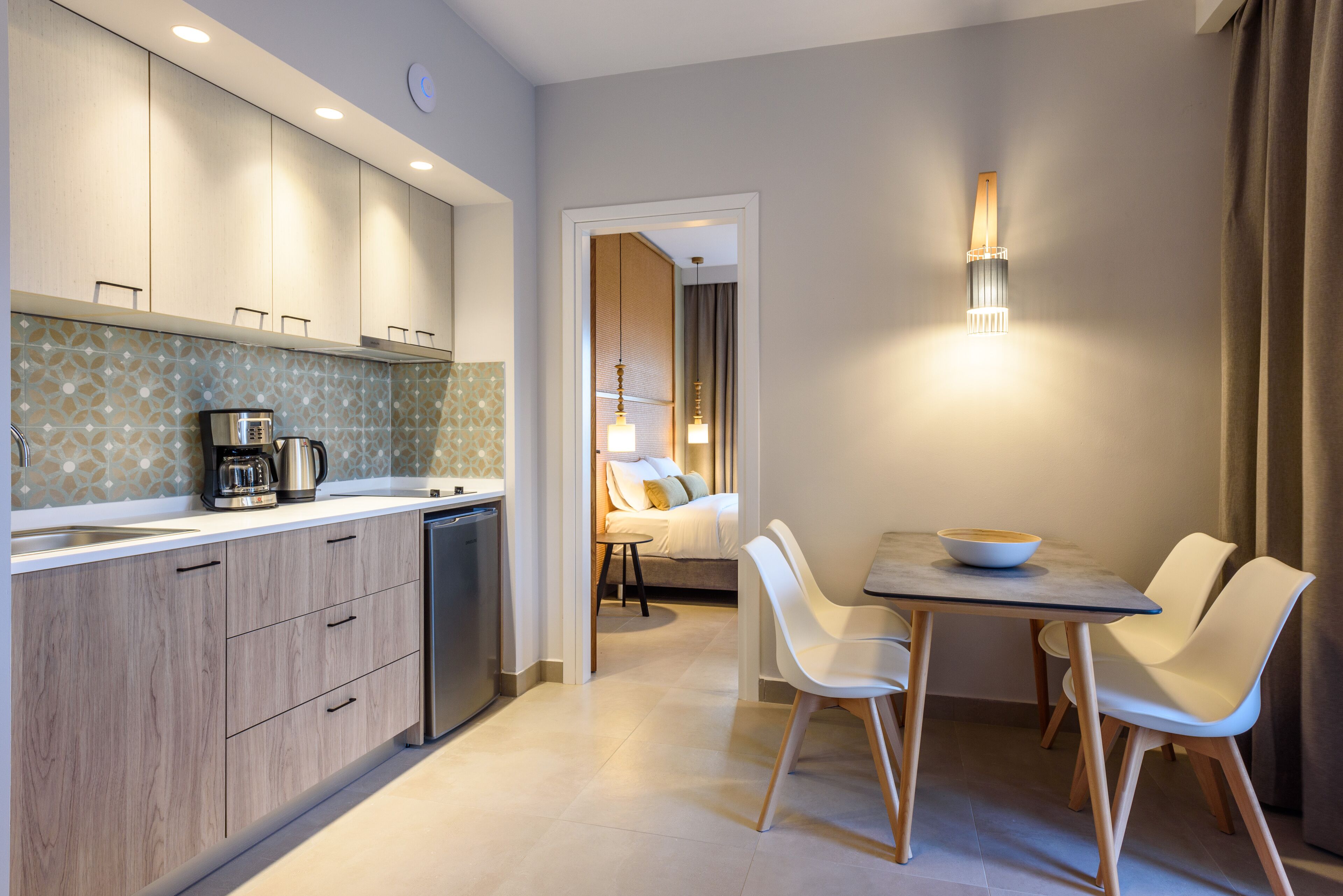 Una cucina moderna con mobili in legno, una macchina da caffè e uno schienale piastrellato. C'è una zona pranzo con tavolo e sedie, e un letto sullo sfondo.