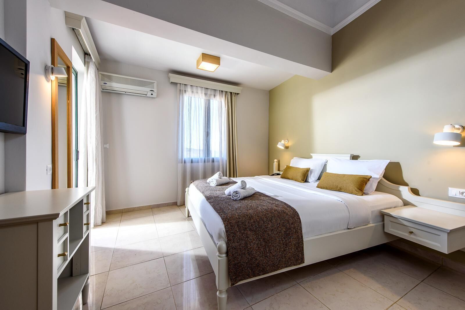 A hotel room with a bed, bedside tables, a TV, and an air conditioning unit.