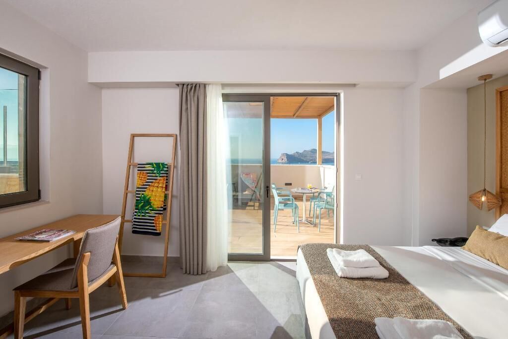 Habitación de hotel con cama, escritorio, silla, balcón con mesa y sillas, y vista al mar.