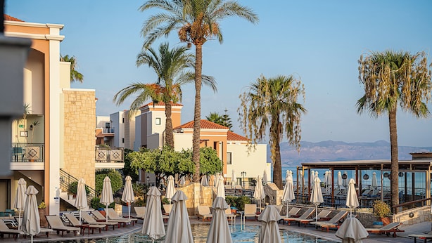A poolside area with lounge chairs and umbrellas, surrounded by palm trees and buildings with balconies. - Caldera Village (Chania)