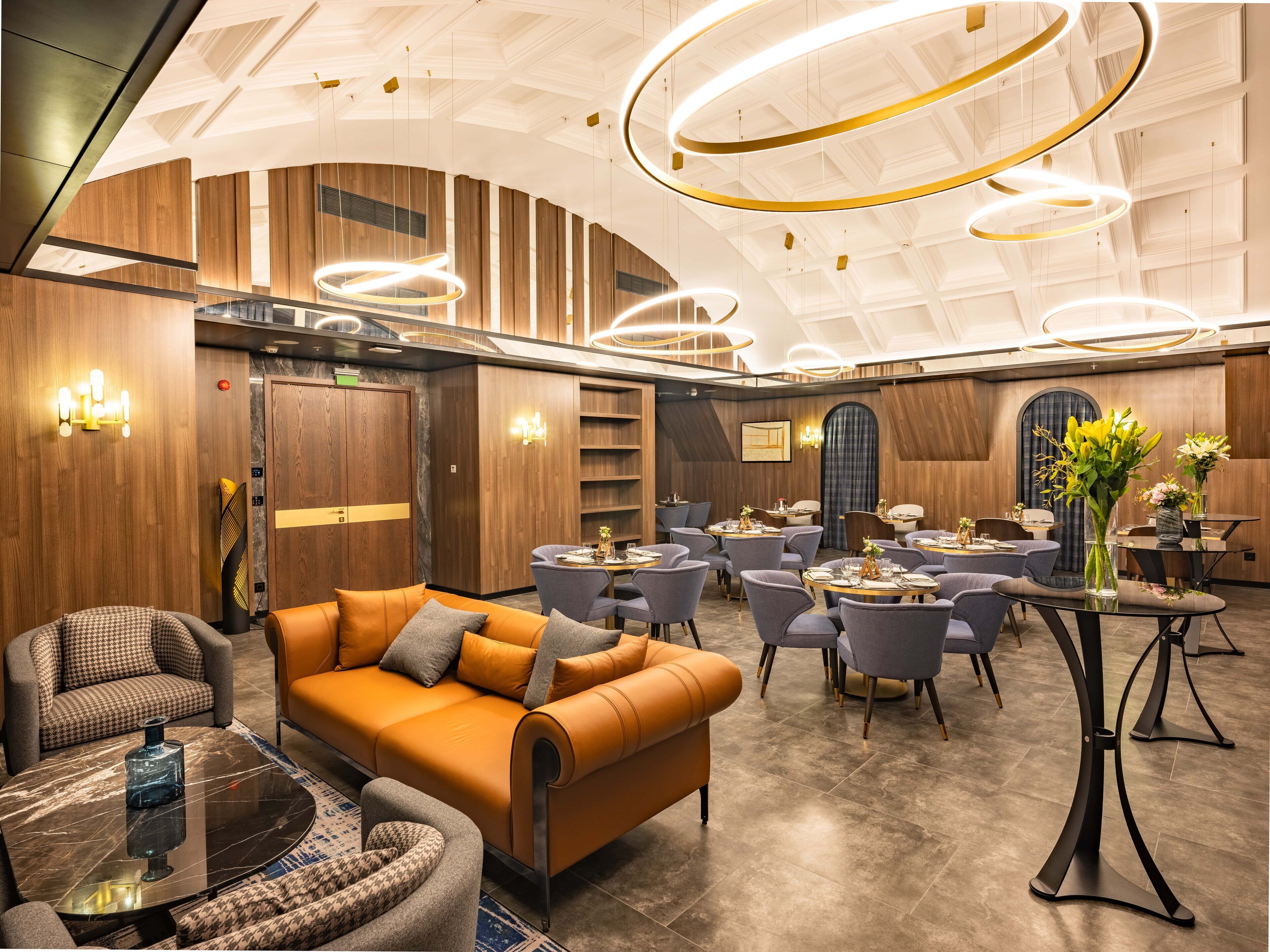 Un moderno vestíbulo de hotel con un área de estar central, una recepción y un comedor con mesas y sillas redondas.