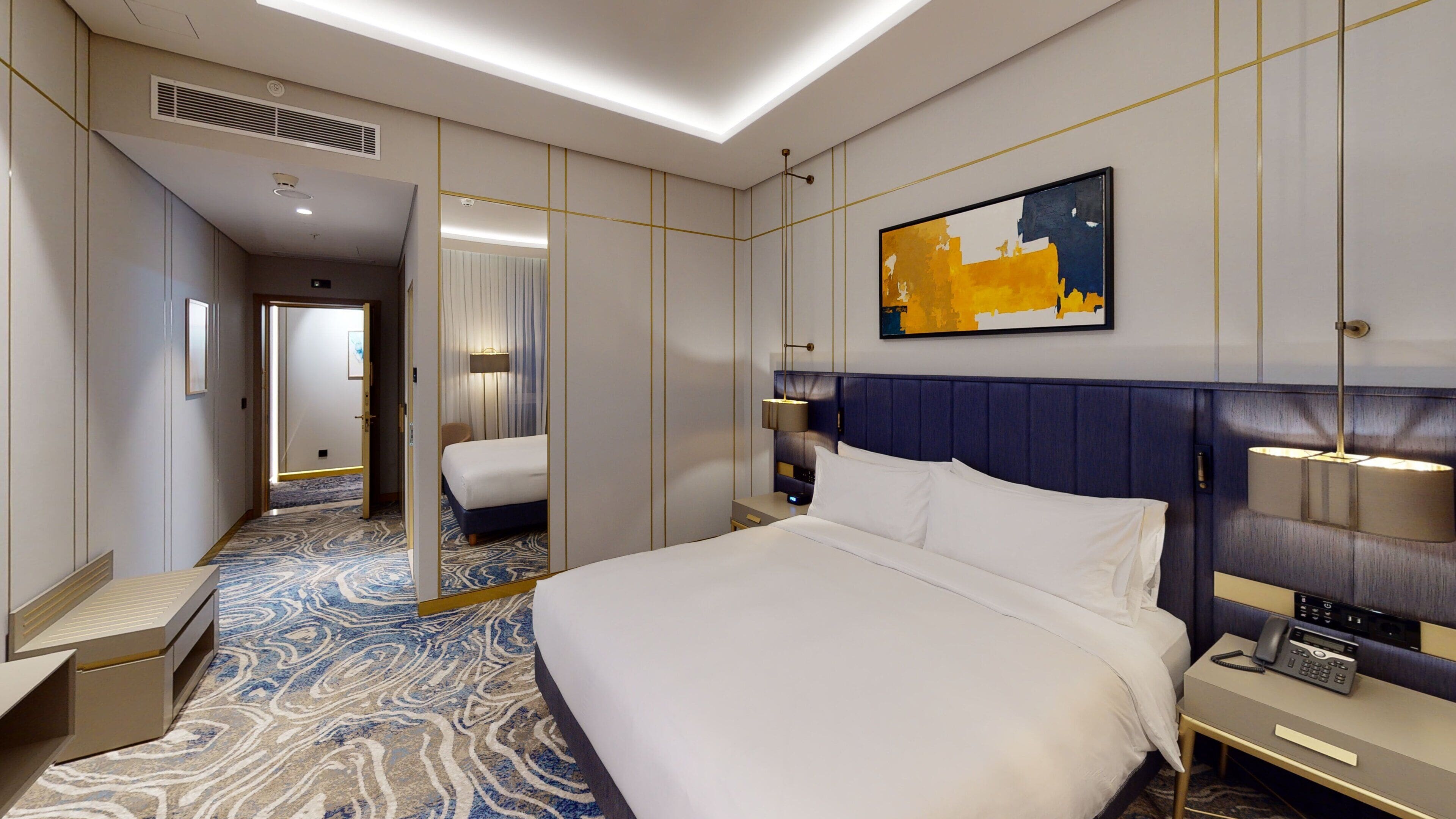 A hotel room with a large bed, bedside tables, a mirror, a painting, and a carpeted floor.