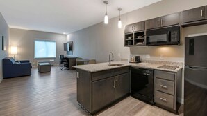 A modern kitchen with dark cabinetry, a built-in microwave, and a dishwasher. There is a dining area with a table and chairs, and a large window providing natural light. - MainStay Suites Cincinnati University - Uptown (Cincinnati)
