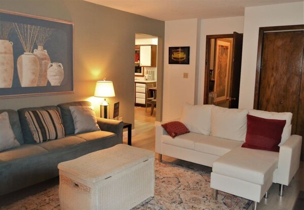 A living room with a sofa, a white armchair, a small table, and a lamp. There is a door leading to another room.