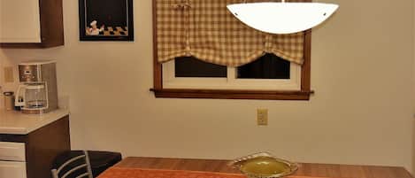 A dining area with a table, chairs, a coffee maker, a checkered window valance, and a wall-mounted menu board.