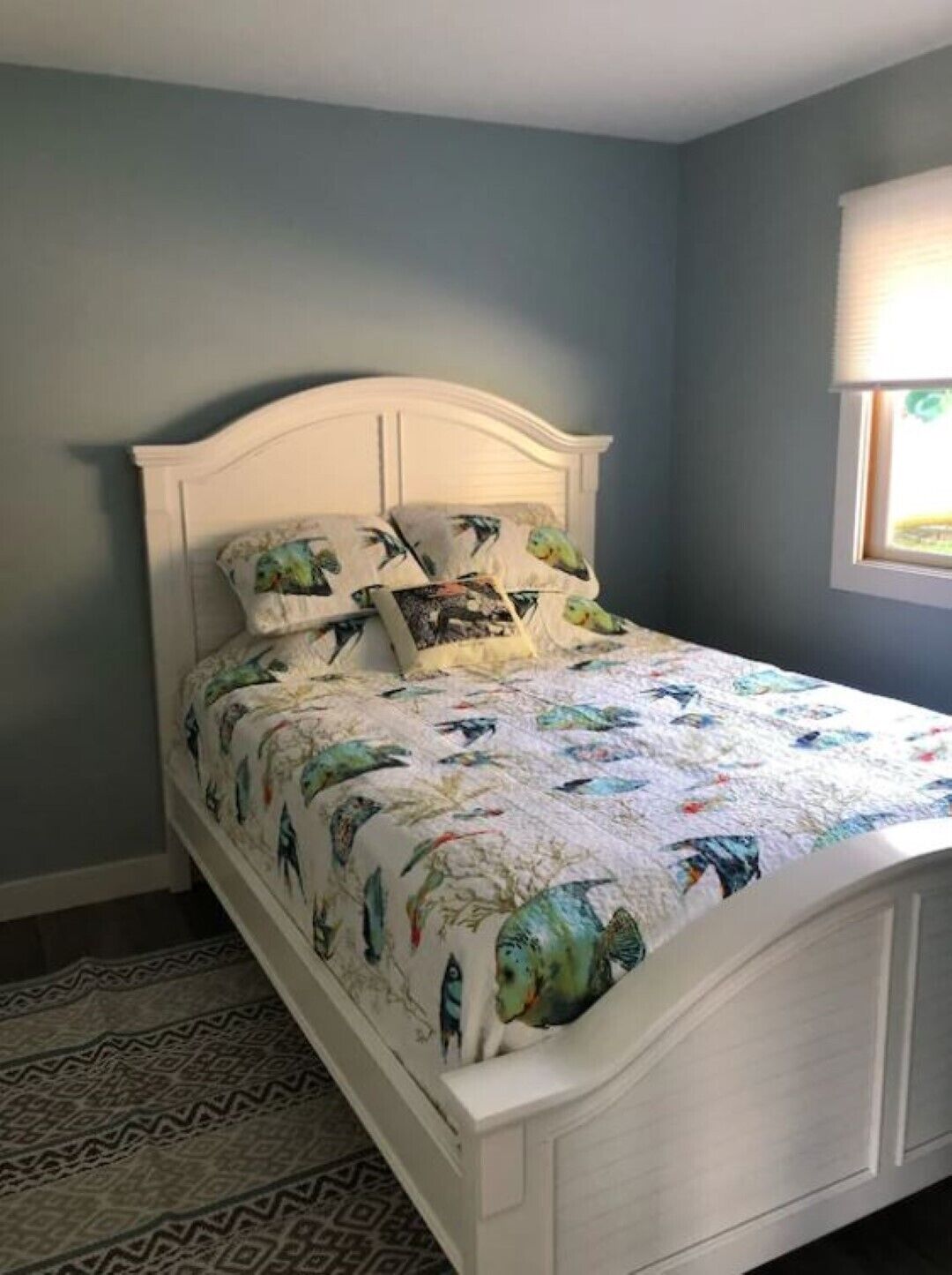 Una cama con cabecera blanca y colcha estampada, una alfombra con diseño y una ventana con persianas.