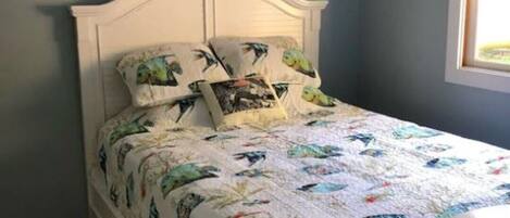 Una cama con cabecera blanca y colcha estampada, una alfombra con diseño y una ventana con persianas.