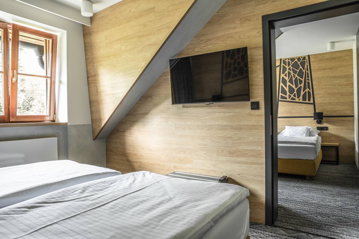 A modern hotel room with a sloped ceiling, a flat-screen TV mounted on the wall, a bed with white bedding, and a view through an open door to another room.