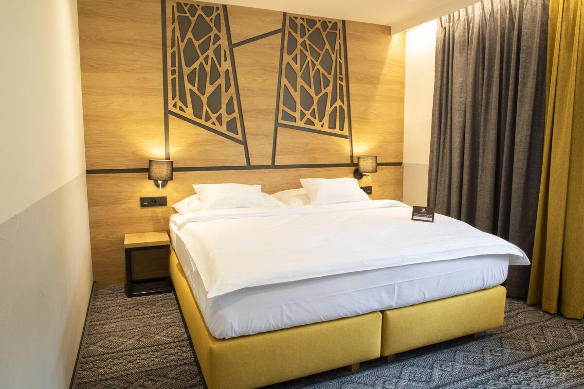 A hotel room with a large bed, a nightstand, a wall-mounted lamp, and a curtain.