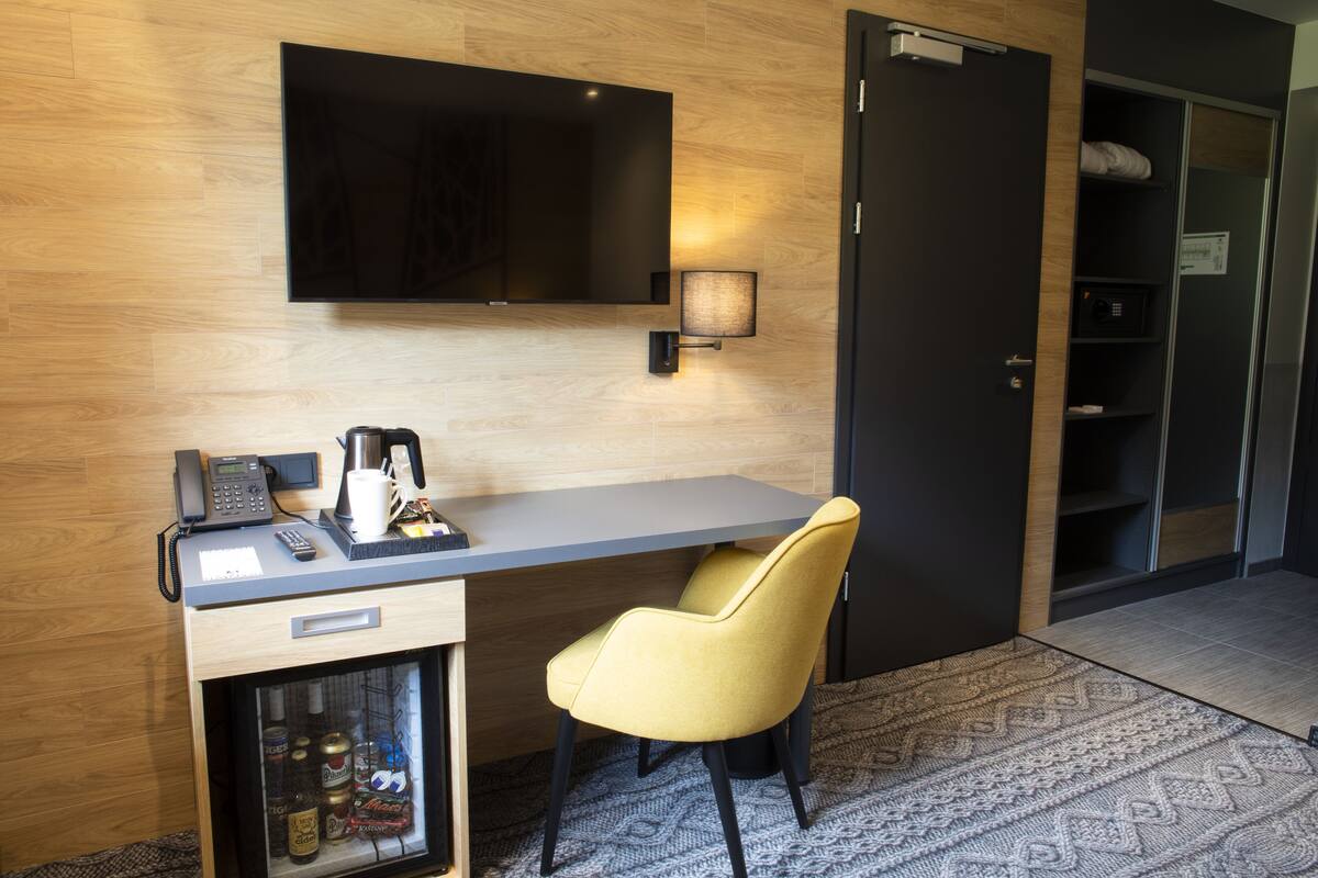 A hotel room with a flat-screen TV mounted on the wall, a desk with a telephone and a coffee maker, a yellow chair, and a minibar with beverages.