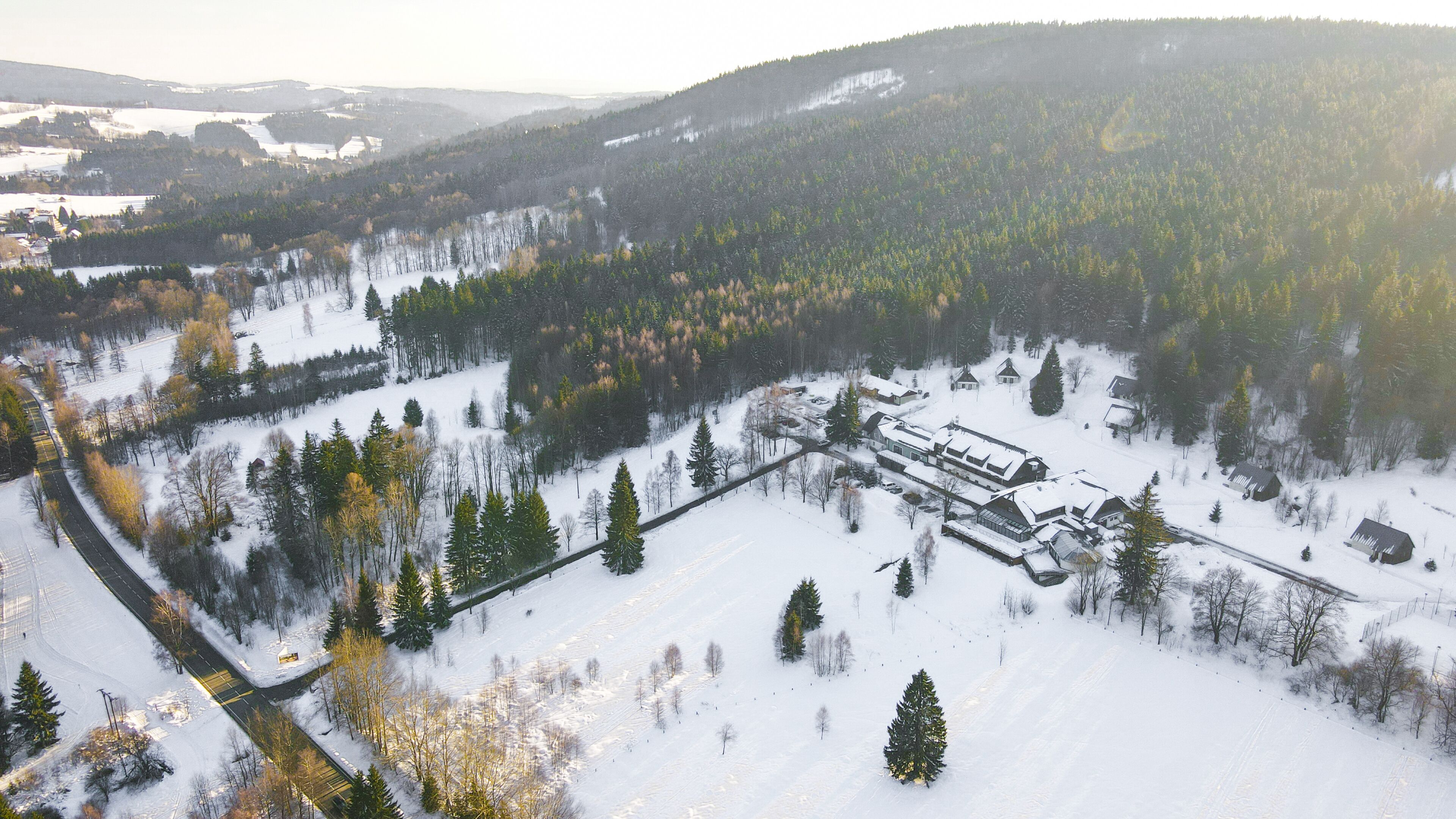 An aerial view of a snow-covered landscape with a cluster of buildings, likely a resort or lodge, surrounded by dense forests and snow-covered fields.