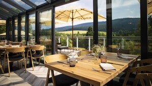 Un área de comedor al aire libre con mesas y sillas de madera, grandes sombrillas y una vista panorámica de montañas y vegetación a través de amplias ventanas.