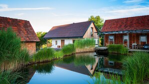 A riverside building with a wooden exterior and a red roof, surrounded by lush greenery and a calm water body.