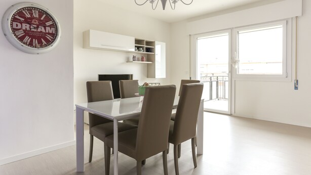 A dining area with a white table and four chairs, a wall-mounted clock, a chandelier, and a window with a view. - Chic Aprtment 10 min to Venice (Mestre)