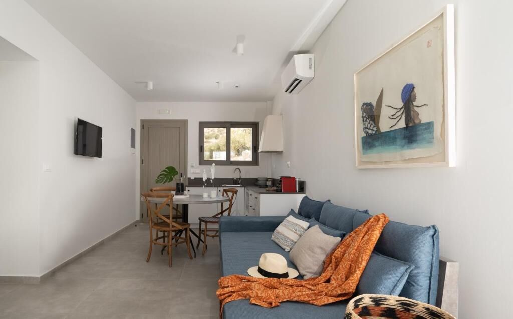 A modern apartment with a living room, dining area, and kitchenette. There is a blue sofa with a hat and a blanket, a dining table with chairs, and a TV mounted on the wall.