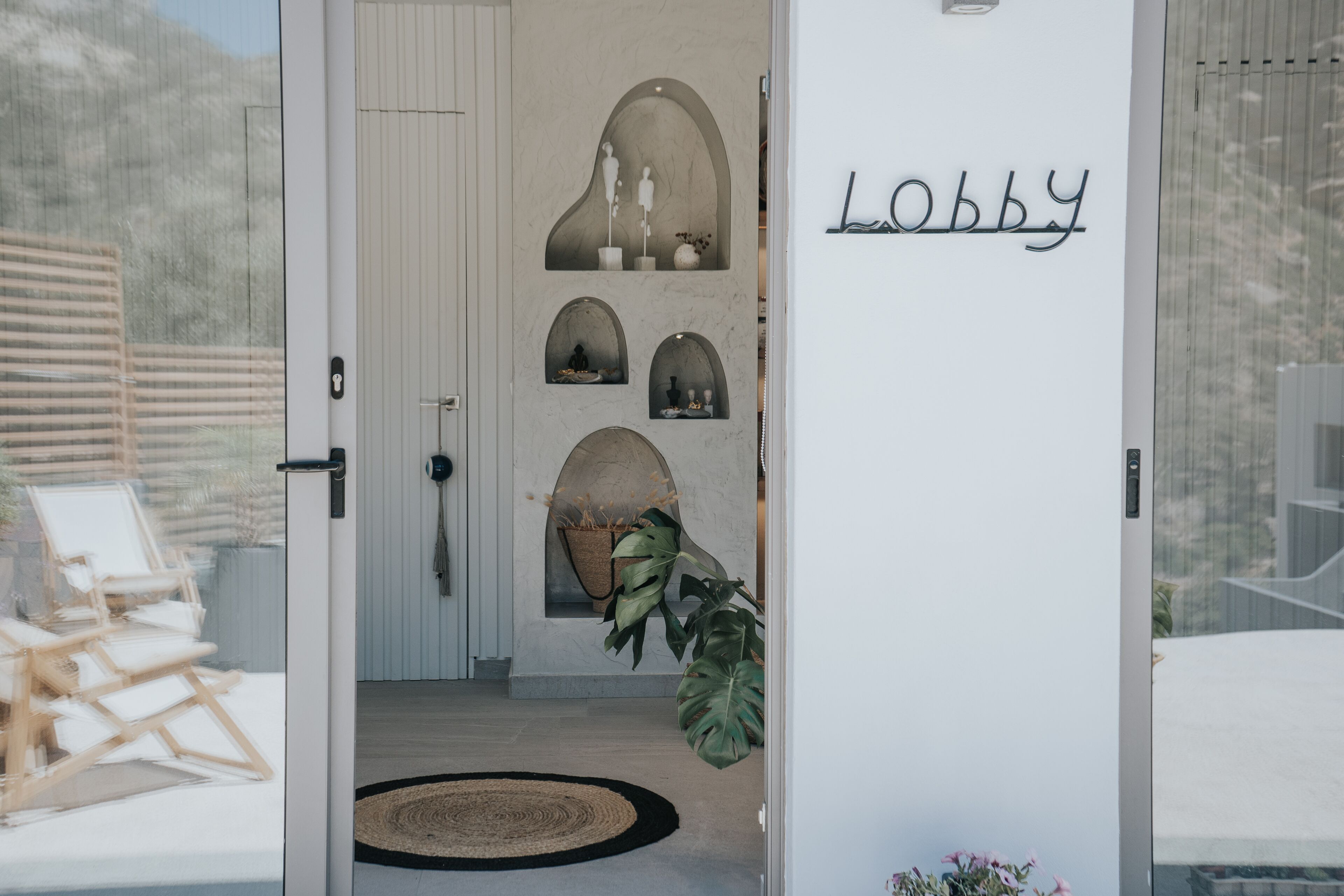 A lobby with a door labeled "Lobby," a white wall with decorative niches, a large plant, and a wooden chair outside.