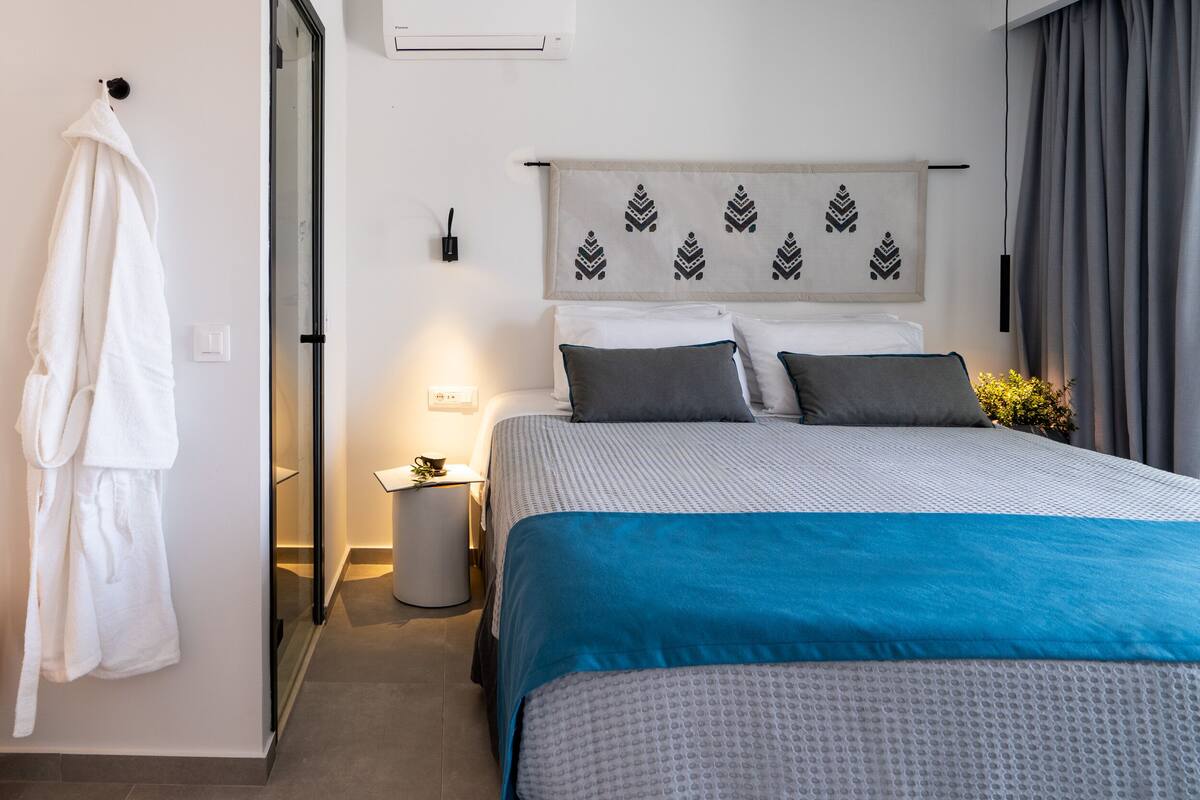 A hotel room with a bed, a robe hanging on the wall, a bedside table with a lamp, and a wall-mounted air conditioner.