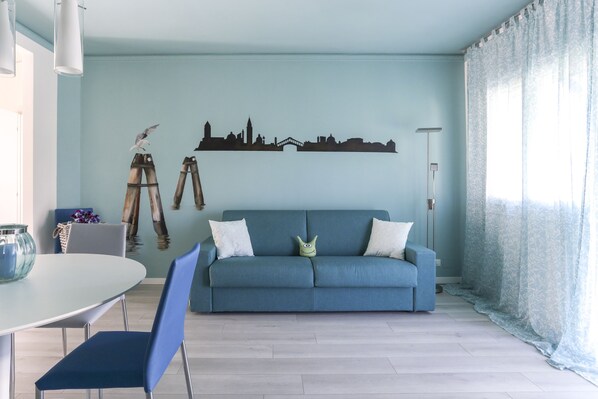 Een woonkamer met een blauwe bank, een eettafel met stoelen en een muurschildering van een stadsgezicht.