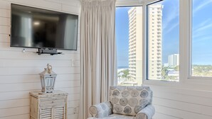 Een hotelkamer met een grote televisie aan de muur, een gestreept fauteuil, een klein bijzettafeltje met een lantaarn, en een uitzicht op hoge gebouwen door een raam.