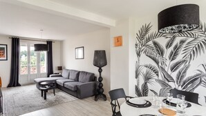 En moderne stue med en grå sofa, et spisebord dækket til fire personer og et stort, sort-hvidt tropisk plantemotiv på væggen.