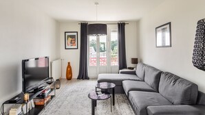 En moderne stue med en grå sofa, et fladskærms-tv, et sofabord og et stort vindue med gardiner.