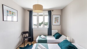 Et moderne hotelværelse med to senge, en stol, et stort vindue med gardiner og kunst på væggene.