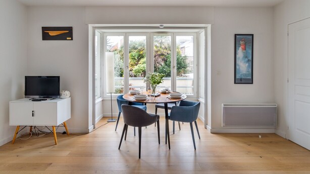A dining area with a round table, chairs, a television on a cabinet, and a large window with a view of greenery. - L'Astrolabe - One Bedroom Apartment, Sleeps 4 (Dinard)