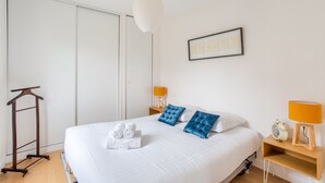 A hotel room with a large bed, two bedside lamps, a bedside table with a clock, and a hanging light fixture. - L'Astrolabe - One Bedroom Apartment, Sleeps 4 (Dinard)