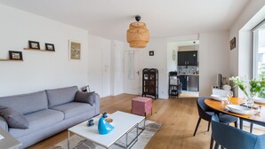 A modern living room with a grey sofa, a white coffee table, and a dining area with a wooden table and chairs. There is a kitchen visible in the background with dark cabinets and a black refrigerator. - L'Astrolabe - One Bedroom Apartment, Sleeps 4 (Dinard)