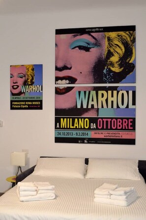A hotel room with a bed, pillows, and folded towels. There are two posters on the wall, one of Marilyn Monroe and another of Warhol's 'A MILANO DA OTTOBRE'. - New "Orange" studio apartment in Sauro (Milano)