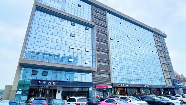 A multi-story hotel with a modern glass facade, several ground-level retail spaces, and a parking lot with cars. - Ji Hotel(shanghai Hongqiao Trans Hub Xujing Branch (Shanghai)