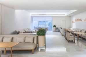 Ein modernes, helles Café mit hoher Decke, großen Fenstern und vielfältigen Sitzgelegenheiten, darunter Sofas und Tische.