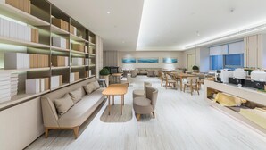 A modern hotel lobby with a sofa, tables, chairs, a coffee station, and a bookshelf. - Ji Hotel(shanghai Hongqiao Trans Hub Xujing Branch (Shanghai)