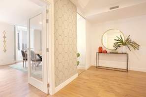Un intérieur moderne avec un plancher en bois, une porte en verre donnant sur un coin repas, un miroir rond posé sur une console et un mur orné d’un motif.