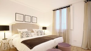 Quarto com uma cama grande, luminárias de cabeceira, uma mesinha e uma janela com cortinas.