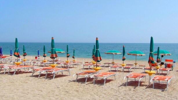 Ein Strand mit Reihen von Liegestühlen und Sonnenschirmen, einem klaren Himmel und dem Meer im Hintergrund.