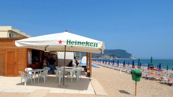 Un bar Heineken situé en bord de plage, avec des tables à l’extérieur, une vue sur la mer et une montagne en arrière-plan.