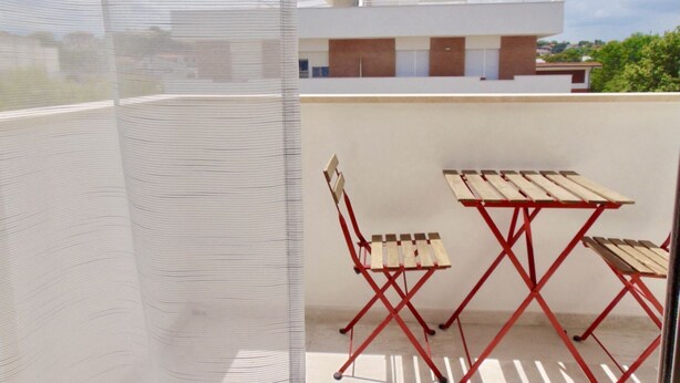 Uma varanda com mesa e cadeiras de metal vermelho, um painel de tela branca e um edifício moderno ao fundo.