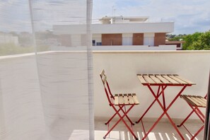 Un balcón con una mesa y sillas de metal rojo, una celosía de malla blanca y un edificio moderno al fondo.