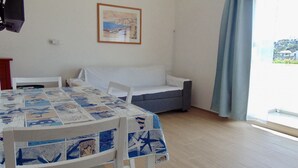 Quarto com mesa de jantar, duas cadeiras brancas, um sofá cinzento, um quadro na parede e vista pela janela com cortinas.