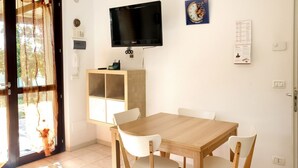 A dining area with a wooden table and white chairs, a TV mounted on the wall, and a cabinet below. - M273 - Marcelli, new three-room apartment with garden (Marcelli)
