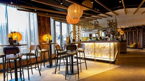 Een moderne bar met hoge krukken en een marmeren toonbank, grote ramen met gordijnen en decoratieve hanglampen.