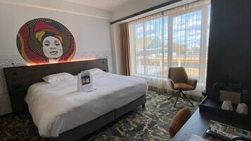 Une chambre d’hôtel comprenant un lit, un bureau avec une chaise, une grande fenêtre avec des rideaux et une œuvre d’art murale.