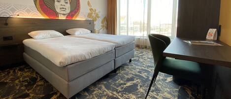 A hotel room with a double bed, a desk, and a chair. There is a large window with curtains and a view of the outside.