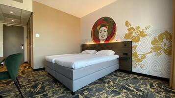 Habitación de hotel con cama, una silla verde, alfombra estampada y una pared con diseño floral.