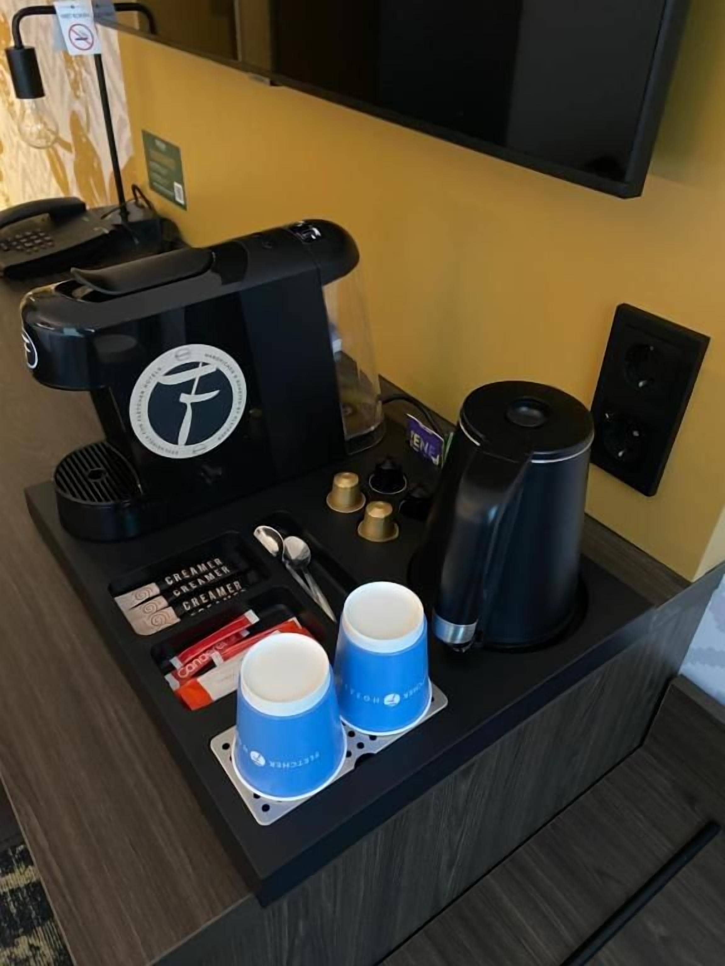 Estación de café en una habitación de hotel, con una cafetera, dos tazas, una cuchara, un sobre de crema y un pequeño letrero.