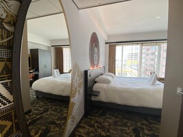 Habitación de hotel con dos camas, un escritorio, una silla y un ventanal con vistas a la ciudad.