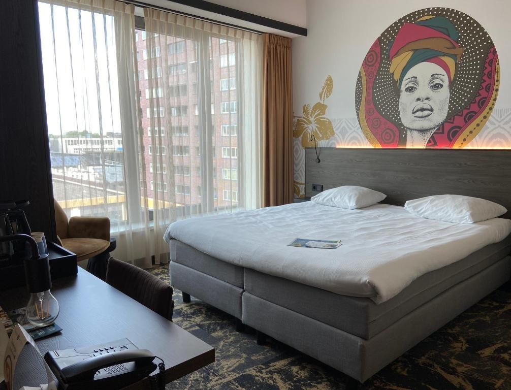 A hotel room with a large bed, a desk with a telephone, and a view of the city through the window.