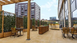 Een terras buiten met houten tafels en stoelen, een houten plantenbak in het midden en een hoge gebouw op de achtergrond.