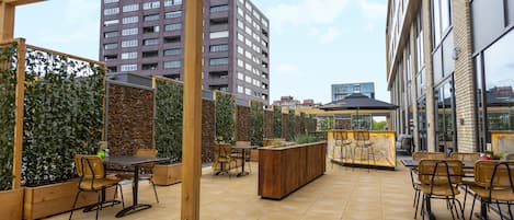 Un espace extérieur aménagé en terrasse, avec des tables et des chaises en bois, un grand bac à fleurs en bois au centre, et un immeuble imposant en arrière-plan.